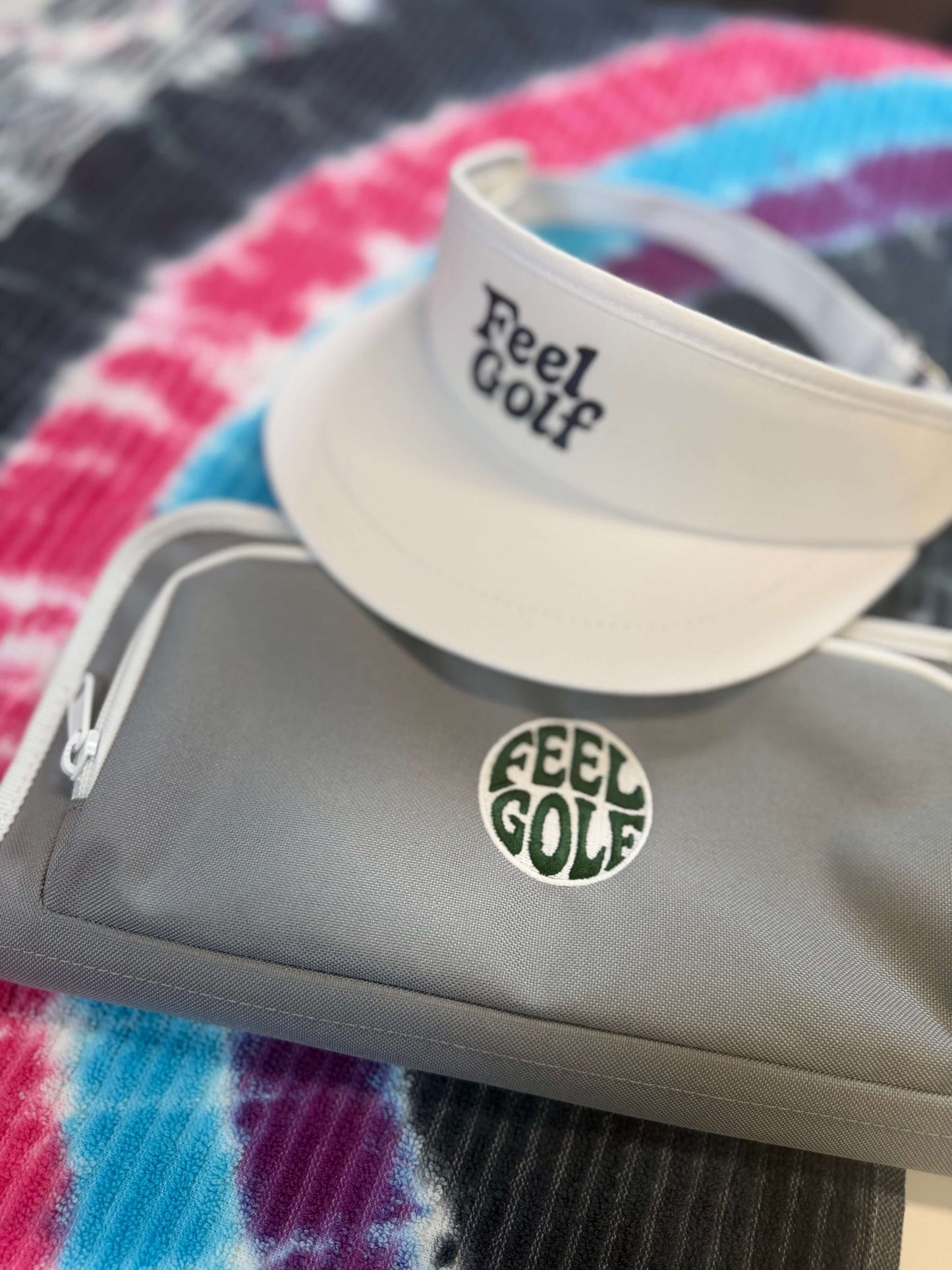 A white visor with 'Feel Golf' printed on it rests on top of a gray zippered pouch with a matching circular 'Feel Golf' logo, placed on a colorful tie-dye blanket.