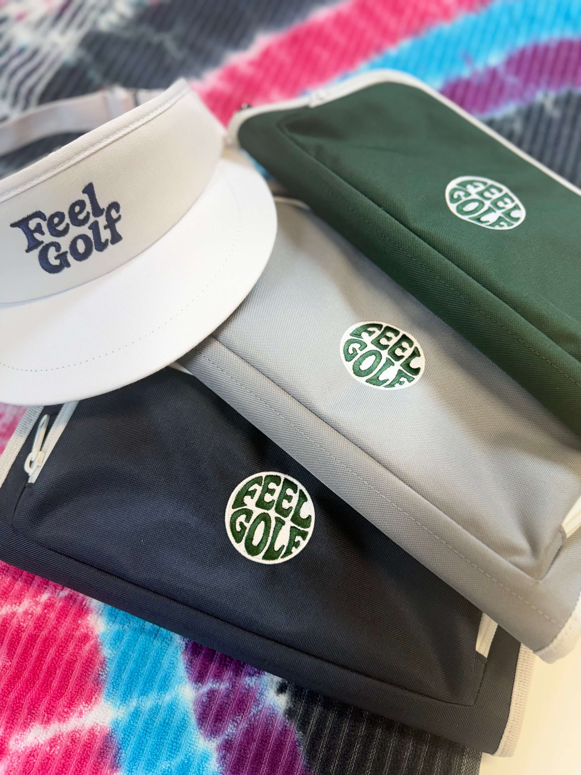 A white visor and three golf organizer pouches, featuring the 'Feel Golf' logo. Jolly Golf bag organizers in grey, dark green, and navy, each featuring the 'Feel Golf' logo, arranged on a colorful tie-dye blanket.
