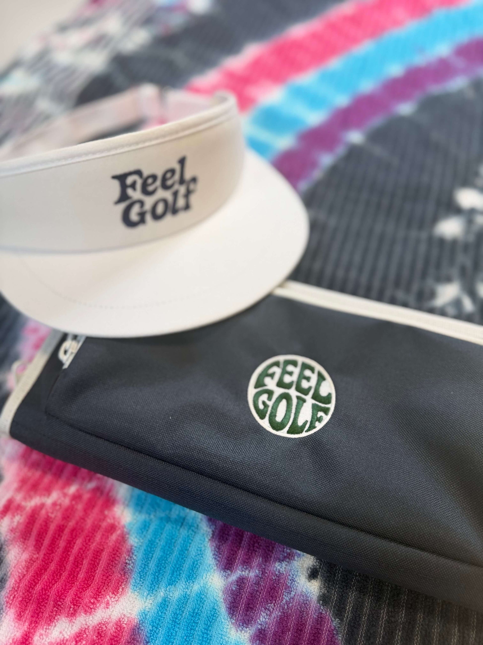 A white visor and a navy golf pouch, both featuring the 'Feel Golf' logo, displayed on a colorful tie-dye blanket.