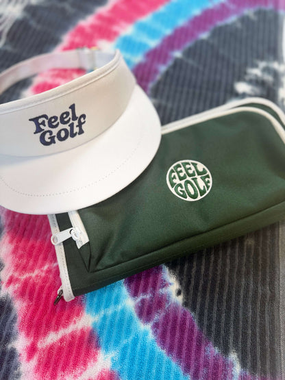 A white visor with 'Feel Golf' printed in navy blue and a dark green zippered Jolly Golf organizer pouch with a white 'Feel Golf' circular logo, placed on a colorful tie-dye blanket.